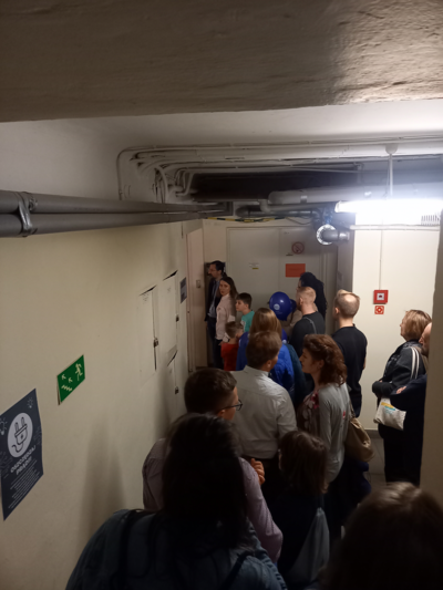  Na zdjęciu fragment korytarza w podziemiach Głównego Urzędu Miar w którym stoi w kolejce spora grupa osób w oczekiwaniu na wejście do laboratorium.
