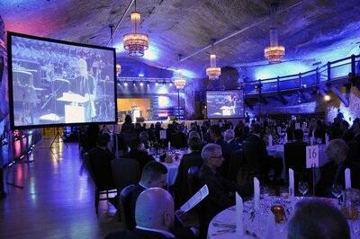  Sala balowa w kopalni soli, przy stolikach siedzą eleganccy goście, w tle widać ekran dużego wyświetlacza.