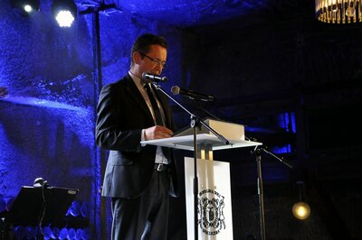  Mężczyzna w garniturze, z ciemnymi włosami i w okularach stoi przed mównicą i mikrofonem.