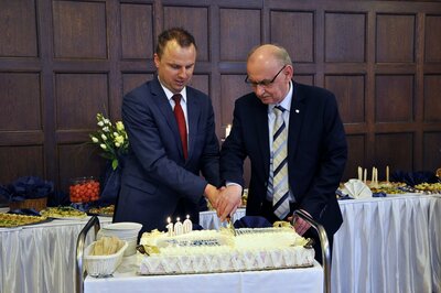  Dwóch mężczyzn w garniturach kroi tort. Za nimi widać na stołach poczęstunek od cateringu.