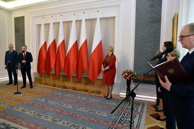  Kobieta - blondynka ubrana na czerwono - stoi na dywanie, a obok niej w wazonie bukiet kwiatów. Za nią rząd biało-czerwonych flag. Z lewej strony stoi dwóch mężczyzn w garniturach. Z prawej strony widać trzy kobiety i mężczyznę, który trzymając w ręku brązową teczkę, stoi przy mikrofonie.