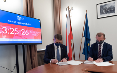  Dwóch mężczyzn w garniturach i maseczkach siedzi przy stole i podpisuje dokumenty. Z lewej strony widać monitor z wyświetlonym czasem urzędowym, nadzorowanym w GUM. Za mężczyznami flagi Polski i Unii Europejskiej, na ścianie stare zdjęcie.