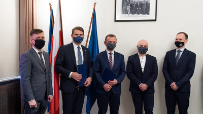  Na zdjęciu widać pięciu stojących mężczyzn w garniturach i maseczkach. Dwóch z nich trzyma w rękach dokumenty. Za mężczyznami flagi Polski i Unii Europejskiej, na ścianie stare zdjęcie.