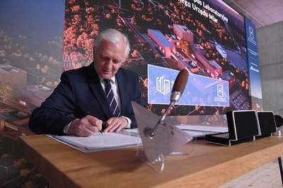  Na zdjęciu mężczyzna z siwymi włosami, w garniturze, podpisuje jakiś dokument przy stole. Z tyłu, za nim duża plansza z wizualizacją Kampusu GUM.