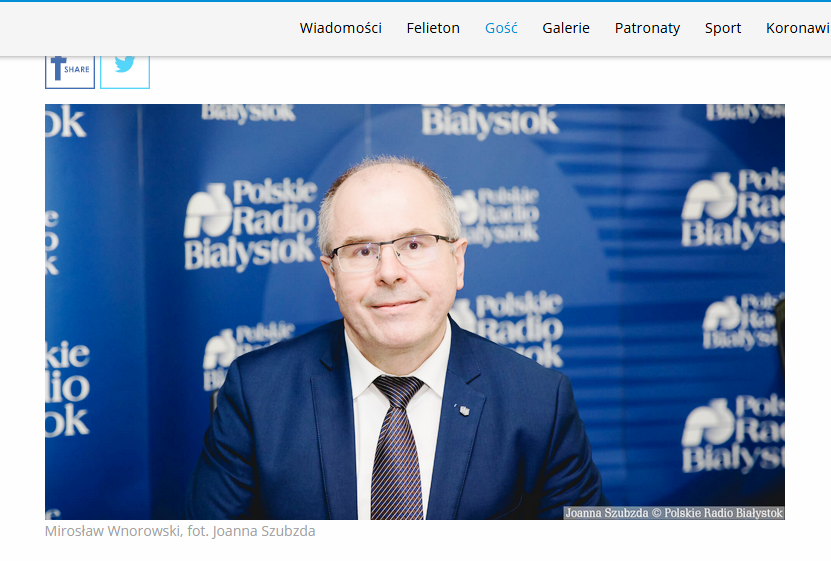 Na zdjęciu uśmiechnięty mężczyzna w okularach, w granatowym garniturze i z krawatem pozuje do zdjęcia na tle logotypów radia Białystok. 