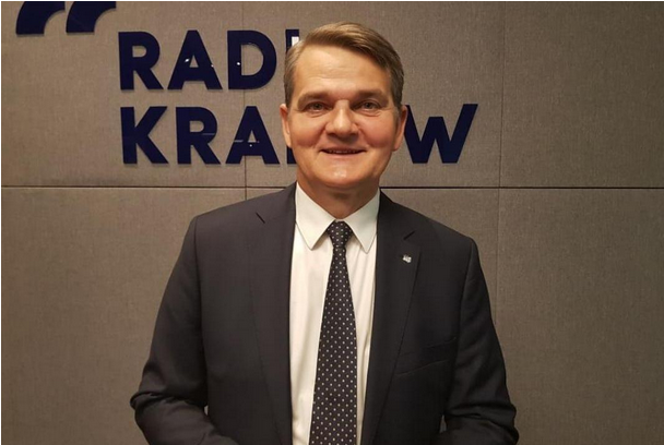 Uśmiechnięty mężczyzna w szarym garniturze stoi na tle ścianki z napisem Radio Kraków
