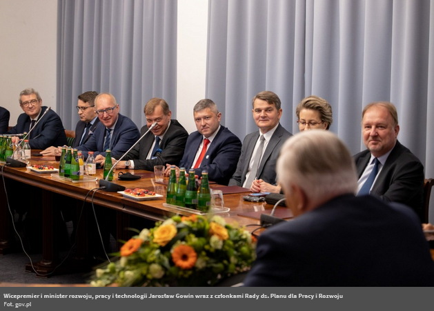 Zdjęcie ze strony Radia Kielce. Za stołem siedzą mężczyźni w garniturach. Spoglądają w kierunku siedzącego tyłem do zdjęcia mężczyzny z siwymi włosami. 